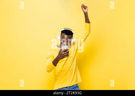 Junger afrikanischer Mann, der auf das Handy blickt und die Hand in einem gelben, isolierten Hintergrund des Studios hebt Stockfoto