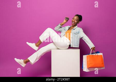 Trendige modische afroamerikanische Frau mit bunten Einkaufstaschen und Kreditkarte, die auf Hocker in rosa Farbe Studio isoliert Hintergrund sitzt Stockfoto