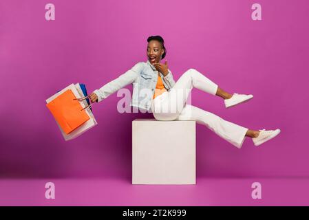 Trendige junge afroamerikanische Frau mit bunten Einkaufstaschen sitzend und blickend zurück in lila rosa isoliertem Studiohintergrund Stockfoto