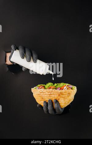 Kocht mit der Hand Soße in Döner-Döner durch gerissene Löcher auf schwarzem Papierhintergrund. Street Food Stockfoto