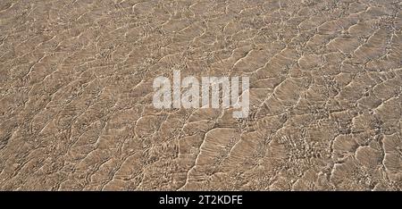 Abstraktes Muster in flachem klarem Wasser über braunem Sand am Strand Stockfoto