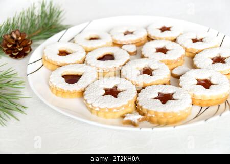 Selbstgemachte Weihnachtskekse mit Marmelade, mit Puderzucker bestreut auf weißem Teller. Weihnachtliche Atmosphäre mit Tannenzweigen und Tannenzapfen Stockfoto