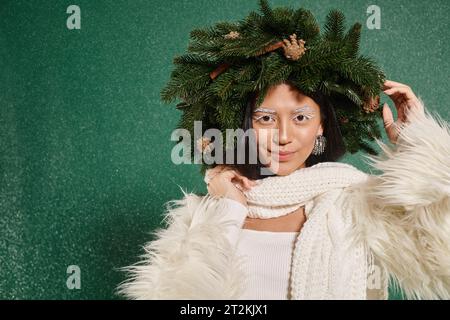 Im Winter, fröhliche Frau mit weißem Make-up und trendigem Outfit, der den Kranz unter fallendem Schnee verstellt Stockfoto