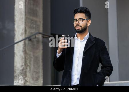 Glücklicher, bärtiger indischer Geschäftsmann, der morgens Kaffee, ein heißes Getränk genießt und draußen lächelt. Entspannen, eine Pause einlegen. Ein arabischer Hindu-Freelancer trinkt Kaffee, um in die Innenstadt zu gehen. Lifestyle Stockfoto