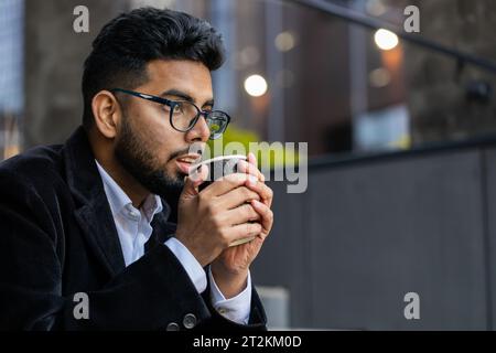 Glücklicher, bärtiger indischer Geschäftsmann, der morgens Kaffee, ein heißes Getränk genießt und draußen lächelt. Entspannen, eine Pause einlegen. Ein arabischer Hindu-Freelancer trinkt Kaffee, um in die Innenstadt zu gehen. Lifestyle Stockfoto