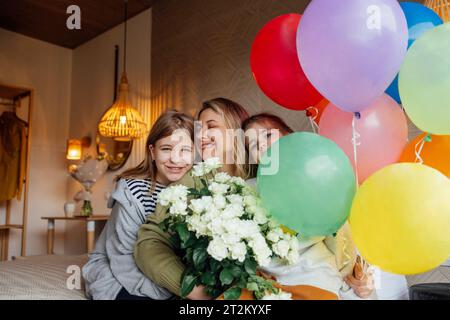 Die lachende Mutter hält Blumenstrauß und bunte Ballons, umarmt ihre Töchter. Junge glückliche Frau feiert ihren Geburtstag zu Hause mit ihrer Familie. Cu Stockfoto