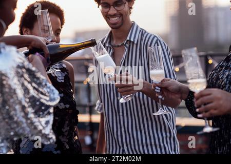 Dunkelhäutiges Mädchen gießt Champagner in Gläser ihrer multinationalen Freunde. Niedliche gemischte Leute, die Spaß auf einer Dachparty haben. Afrikanische Frauen im Eleg Stockfoto