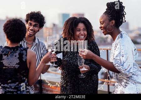 Dunkelhäutiges Mädchen gießt Champagner in Gläser ihrer multinationalen Freunde. Süße, glückliche Leute, die Spaß auf einer Dachparty haben. Afrikanische Frauen in elegantem d Stockfoto