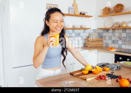 Eine lächelnde, attraktive asiatische Frau trinkt frisch gepressten Orangensaft. Eine charmante junge Koreanerin hält ein Glas köstlichen Saft und lacht in der Th Stockfoto