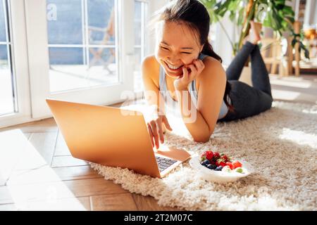 Die junge asiatische Frau liegt auf dem Teppich im Wohnzimmer und benutzt einen Laptop. Die attraktive Koreanerin plaudert mit Freunden und lächelt auf dem Computerbildschirm. Lau Stockfoto