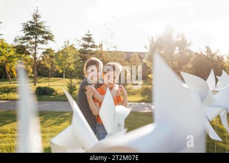 Zwei süße Kinder kuscheln sich im Hinterhof des Hauses. Bruder und Schwester in der Nähe der weißen Windturbinen. Ein Junge und ein Mädchen haben Spaß auf einem grünen Rasen draußen Stockfoto