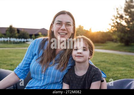 Schöne junge Frau mit Brille umarmt draußen ihre süße Tochter. Die charmante brünette Mutter und ihr kleines Mädchen in lässigen Klamotten lächeln im Backyar Stockfoto