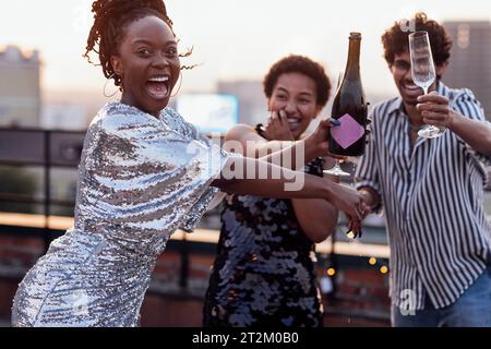 Dunkelhäutiges Mädchen gießt Champagner in Gläser ihrer multinationalen Freunde. Süße, glückliche Leute, die Spaß auf einer Dachparty haben. Afrikanische Frauen in elegantem d Stockfoto