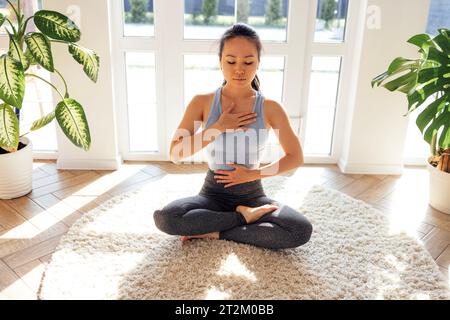 Junge attraktive asiatische Frau sitzt in Lotusposition auf Teppich im Wohnzimmer. Ein charmantes Mädchen im Fitnessanzug meditiert und überwacht ihre Atmung Stockfoto