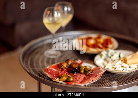 Zwei Gläser kühlen Sekt und Teller mit verschiedenen Snacks auf einem runden Metall-Couchtisch. Weiße Teller mit Schinkenscheiben, Salami, Kapern, Oliven und Stockfoto