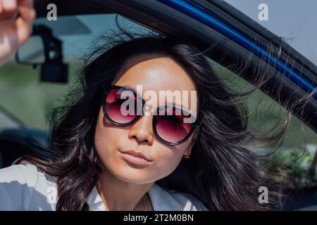 Nahaufnahme eines weiblichen Porträts einer jungen attraktiven asiatischen Frau im Auto. Bezaubernde Koreaner in stilvoller Sonnenbrille im Freien. Das schwarze Haar des Mädchens ist Blowi Stockfoto