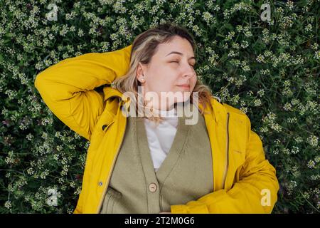 Der Blick von oben auf eine charmante junge Frau hat ihre Augen geschlossen und liegt auf einer blühenden Wiese. Eine attraktive, pralle Blondine in einer gelben Jacke legt ihre Hand Stockfoto