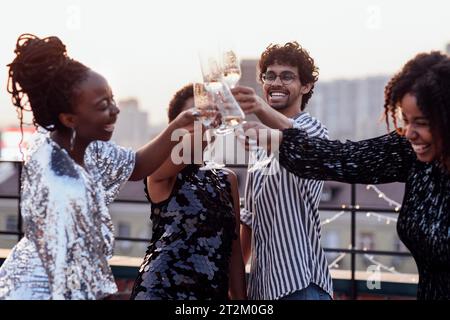 Dunkelhäutiges Mädchen gießt Champagner in Gläser ihrer multinationalen Freunde. Süße, glückliche Leute, die Spaß auf einer Dachparty haben. Afrikanische Frauen in elegantem d Stockfoto