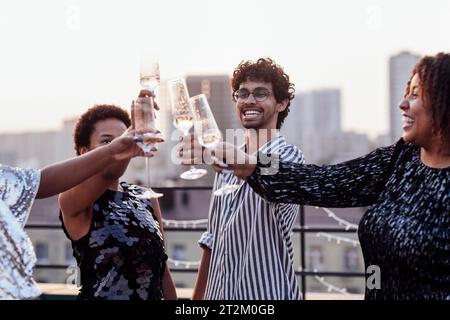 Dunkelhäutiges Mädchen gießt Champagner in Gläser ihrer multinationalen Freunde. Süße, glückliche Leute, die Spaß auf einer Dachparty haben. Afrikanische Frauen in elegantem d Stockfoto