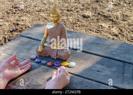 Frauenhände meditieren vor einer Buddha-Statue mit bunten Chakra-Steinen auf einem Holztisch in einem Park in der Sonne Stockfoto