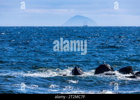 Wellen brechen auf Felsen vor der Küste der Halbinsel Kintyre, mit dem Barquentine „Thalassa“ (erbaut 1980), der sich Aisla Craig, Argyll & Bute nähert. Stockfoto