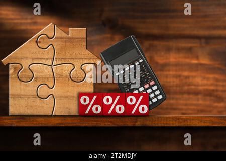 Holzhaus aus Puzzleteilen, Holzblöcken mit roten Prozentzeichen und einem Taschenrechner, auf einem Holzregal oder Tisch mit Kopierraum. Stockfoto