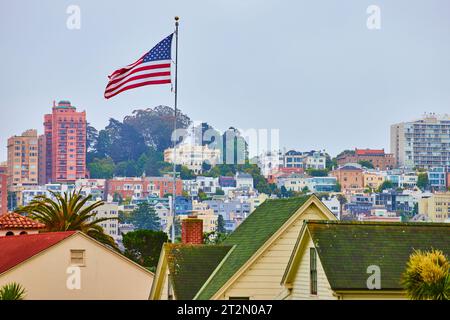 Amerikanische Flagge winkt im Wind über grünen Dächern mit bewölktem Himmel und Stadt dahinter Stockfoto