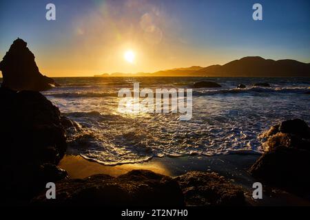 Herrlicher goldener Sonnenuntergang über Meereswellen, die an die Küste mit fernen Bergen heranrollt Stockfoto