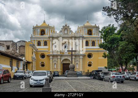 Guatemala, La Antigua - 20. Juli 2023: Klosterkirche Nuestra Senora de la Merced. Gelbe Fassade mit Glockentürmen und Kreuz vorne unter Grau Stockfoto