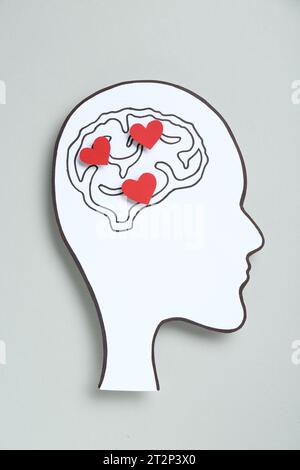 Emotionales Denken. Papierausschnitt des menschlichen Kopfes mit Zeichnung des Gehirns und der roten Herzen auf grauem Hintergrund, Draufsicht Stockfoto