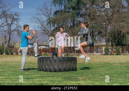 Drei Sportfreunde, die Sprünge auf einem riesigen Lkw-Reifen machen. Sie trainieren im Park, an einem sonnigen Tag Stockfoto