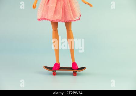 Barbie-Puppe in einem rosa Kleid auf einem Skateboard auf blauem Hintergrund Stockfoto