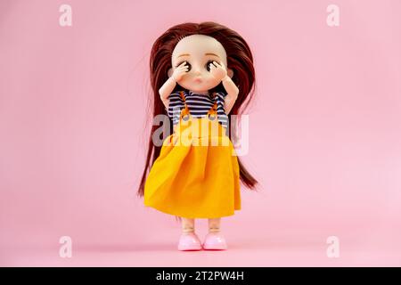 Eine Kinderspielzeugpuppe mit dunklen Haaren in einem orangen Kleid auf rosa Hintergrund. Kinderspielzeug aus Plastik. Puppenspiele für die Fantasie. Der verängstigte d Stockfoto