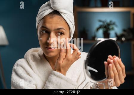 Hispanische Millennial Frau trägt Bademantel und Handtuch im Schlafzimmer Anwendung verjüngende Straffung Antiage Gesichtspflege Creme Behandlung, sitzend mit Stockfoto