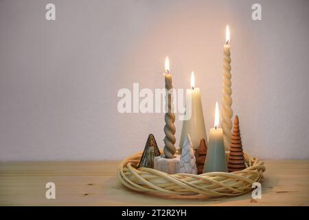 Einfache Adventsdekoration in natürlichen Farben mit verschiedenen Kerzen und kleinen künstlichen Weihnachtsbäumen, die in einem Kranz aus Korbgeflecht auf einem Wald angeordnet sind Stockfoto