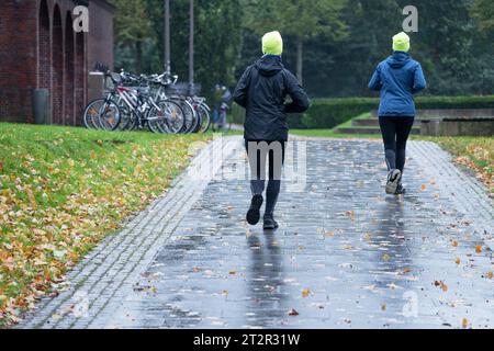 Zwei sportliche Frauen von hinten mit neongelben Mützen joggen im Regen durch einen Herbstpark, es gibt kein schlechtes Wetter für gesunde Outdoor-Action Stockfoto