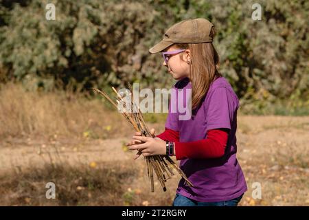 Teenager-Mädchen, in bequemen Anzügen gekleidet, sammelt neugierig Feuerholz für Feuer, während sie in der Natur wandern. Stockfoto