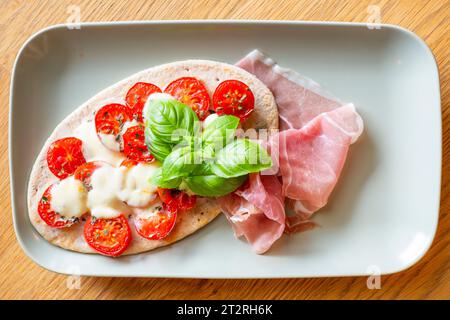 Tomaten-Mozzarella-Pita auf Fladenbrot mit frischem Parmaschinken und Basilikumblättern, serviert auf einem rechteckigen Teller Stockfoto