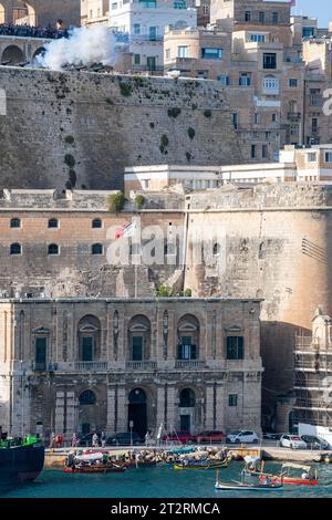 Aus Valletta, der Hauptstadt Maltas, wird eine Kanone abgefeuert, im Vordergrund befinden sich einige traditionelle maltesische Dghajsa (ähnlich einer Gondel), die aus Senglea stammen. Stockfoto