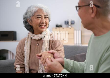 Liebevolles asiatisches Seniorenpaar, das sich auf dem Sofa warm umarmt und ihre dauerhafte Liebe und Zweisamkeit feiert. Ältere Ehemann und Ehefrau sind glücklich Stockfoto