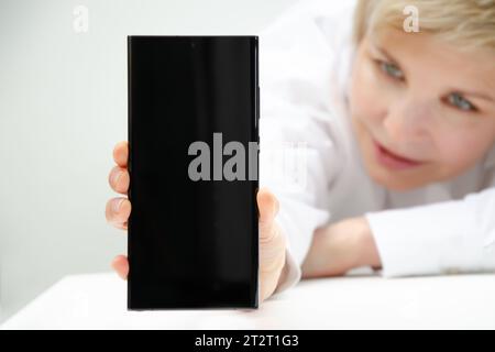 Die Frau hält das Smartphone mit dem Bildschirm nach vorne. Das Gesicht der Frau ist im Hintergrund. Weißer Hintergrund. Kopierbereich. Nahaufnahme eines Smartphone-Scre Stockfoto