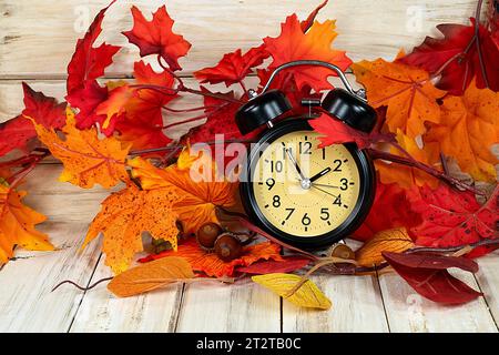 Schwarzer Retro-Wecker in Herbstlaub und Eicheln auf rustikalem Holz zum Zeitwechsel Stockfoto