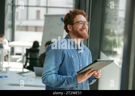 Lächelnder bärtiger Geschäftsmann, der am digitalen Tablet arbeitet, während er im Büro in der Nähe des Fensters steht Stockfoto