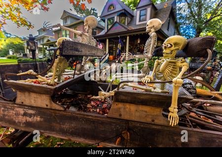 Zwei Plastik-Skelette sitzen eng beieinander als Halloween-Dekorationen auf der Kiste eines geschmückten Pferdewagens im Vorgarten eines viktorianischen Hauses im Central Neighbourhood von Traverse City, Michigan, USA; eines hat den Arm erhoben, das andere hat Kulleraugen aufgeklebt. Stockfoto