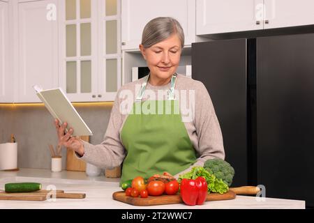 Seniorin mit Rezeptbuch und Produkten am Tisch in der Küche Stockfoto