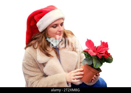 Frau in weihnachtsmann Hut und medizinische Gesichtsmaske wählt weihnachten Poinsettia Blume im Geschäft mit Neujahrsgeschenken, isoliert auf weißem Hintergrund Stockfoto