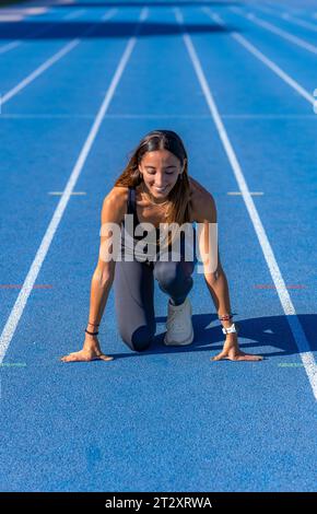 Wunderschönes junges Läufermädchen, gebräunt mit langen dunklen Haaren, lächelnd auf die Rennstrecke an der Startlinie einer blauen Laufstrecke mit den Händen aufliegen Stockfoto