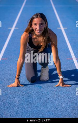 Schöne junge Läuferin, gebräunt mit langen dunklen Haaren, lächelt an der Startlinie einer blauen Leichtathletikbahn und blickt mit gerissenen Händen in die Kamera Stockfoto