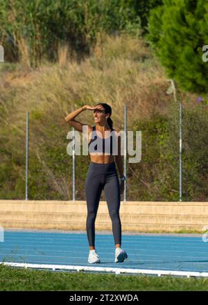 Schöne junge gebräunte Frau mit straffem athletischem Körper, gekleidet in einem Oberteil und enge Sportswear, die glücklich lächelt und ihre Augen vor der Sonne schützt Stockfoto