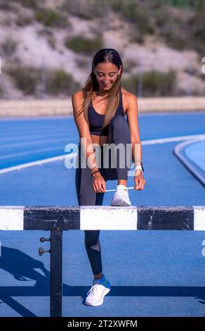 Schönes junges Läufermädchen, gebräunt mit langen braunen Haaren, lächelt mit gebeugtem Bein, lehnt sich auf einem Hindernis auf einer Laufbahn, bindet ihre Schnürsenkel Stockfoto